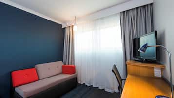 A hotel room with a sofa, a television on a wooden stand, and a window with curtains.
