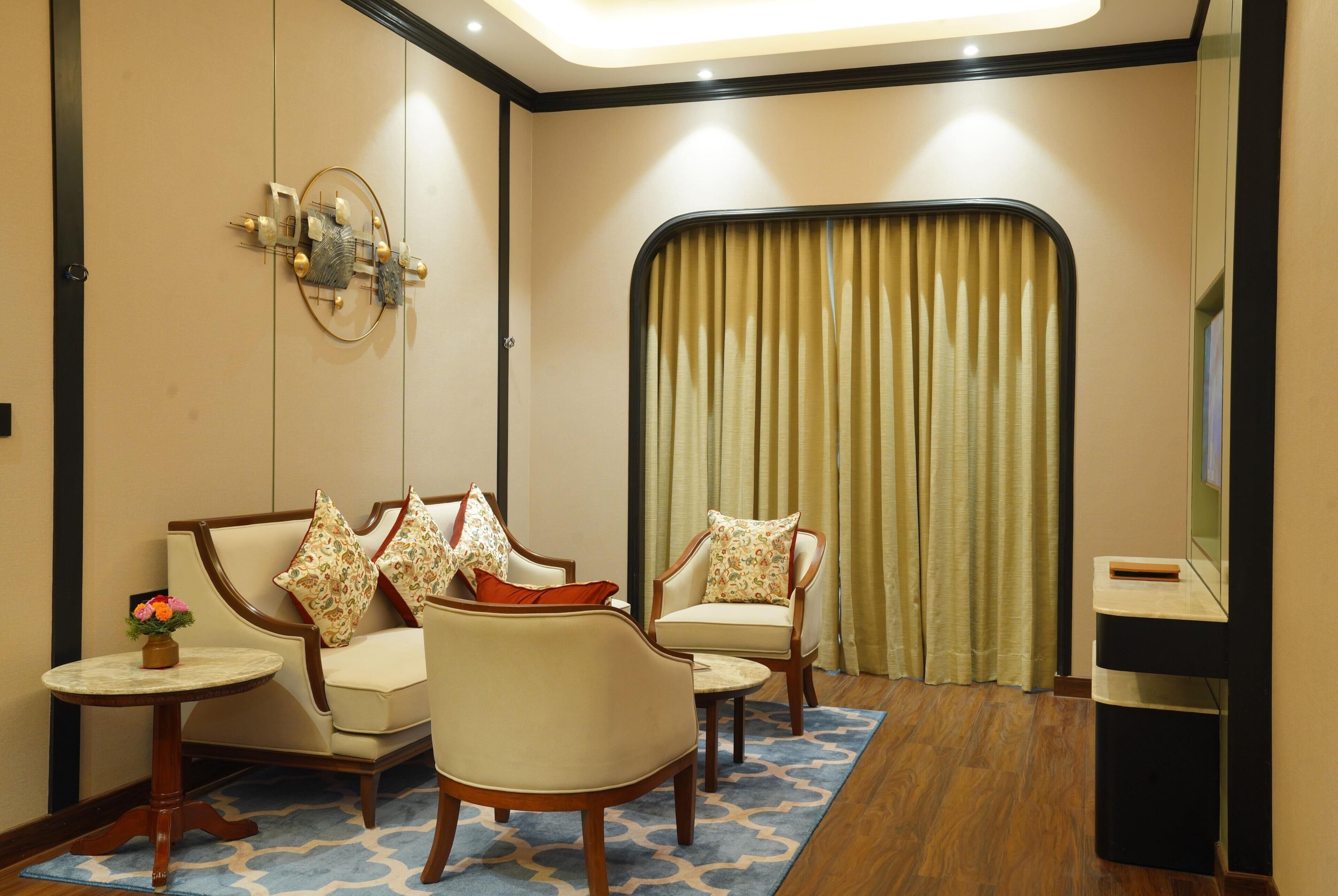 Quarto de hotel com sofá, duas poltronas, uma mesinha de centro com um vaso, um espelho e uma televisão.