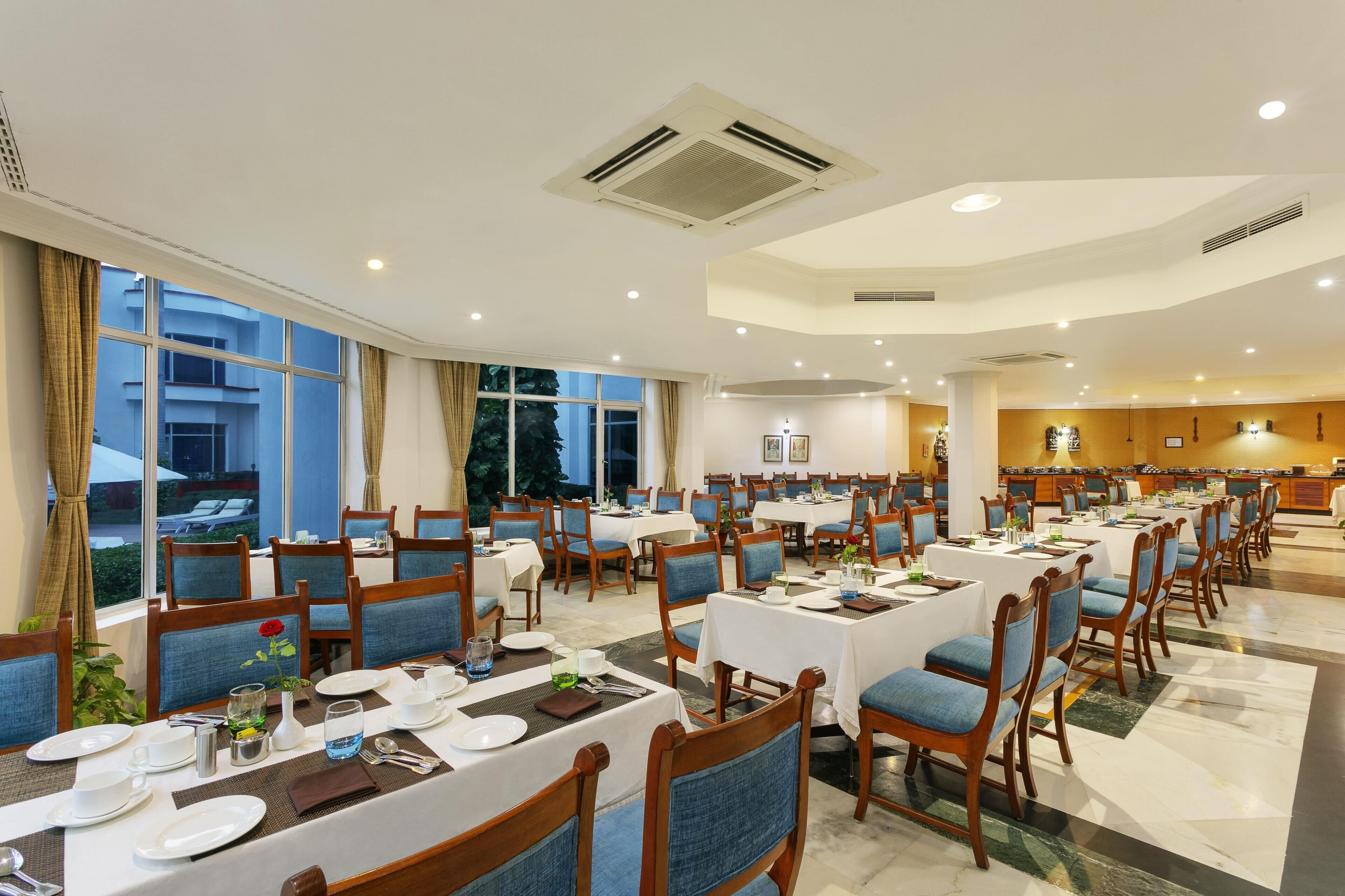 A hotel dining area with multiple tables and chairs, large windows, and a kitchen in the background.