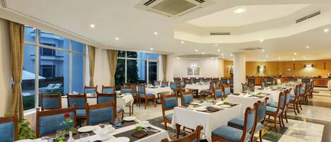 A hotel dining area with multiple tables and chairs, large windows, and a kitchen in the background.