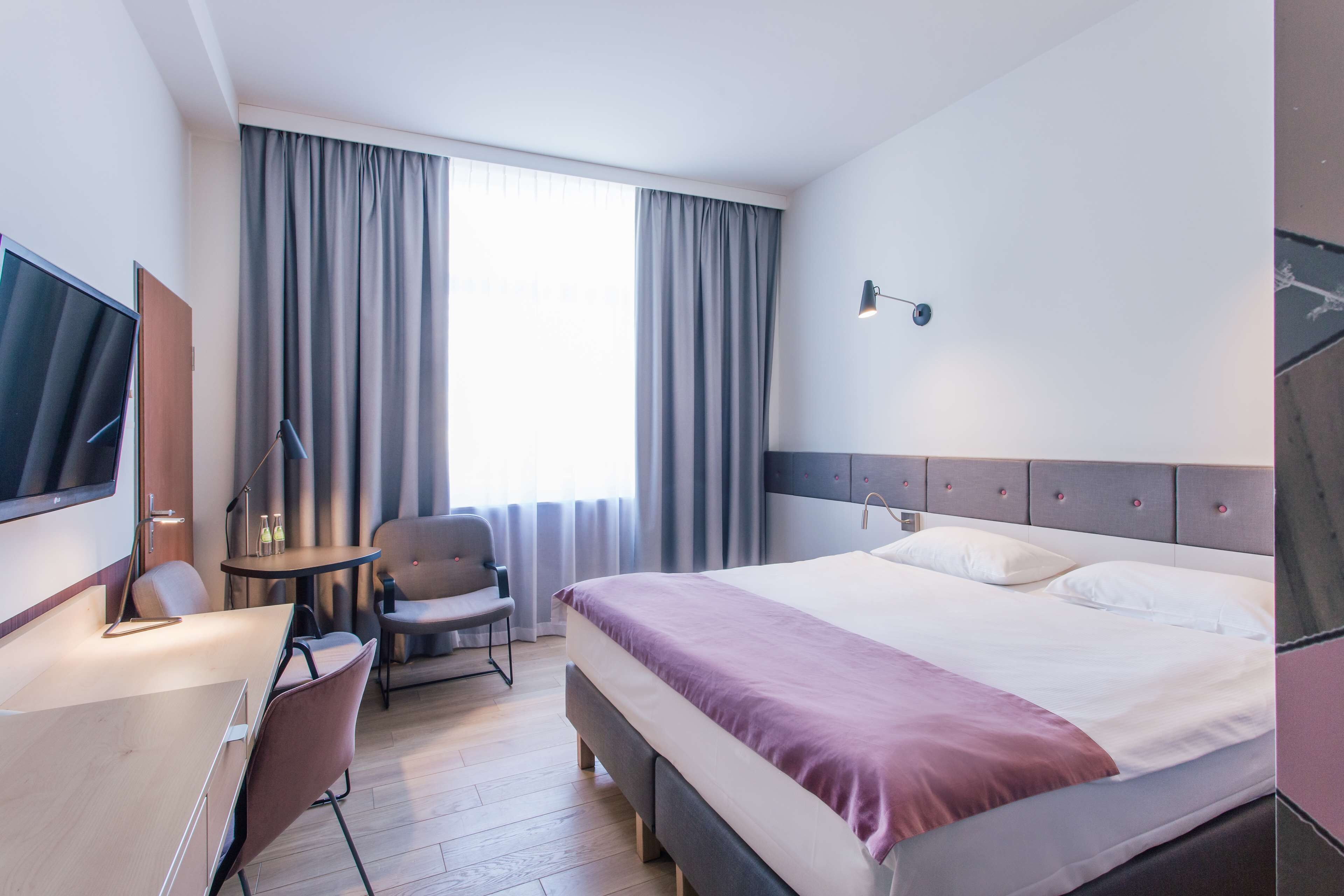 A hotel room with a bed, a desk, a chair, a TV, and a window with curtains.
