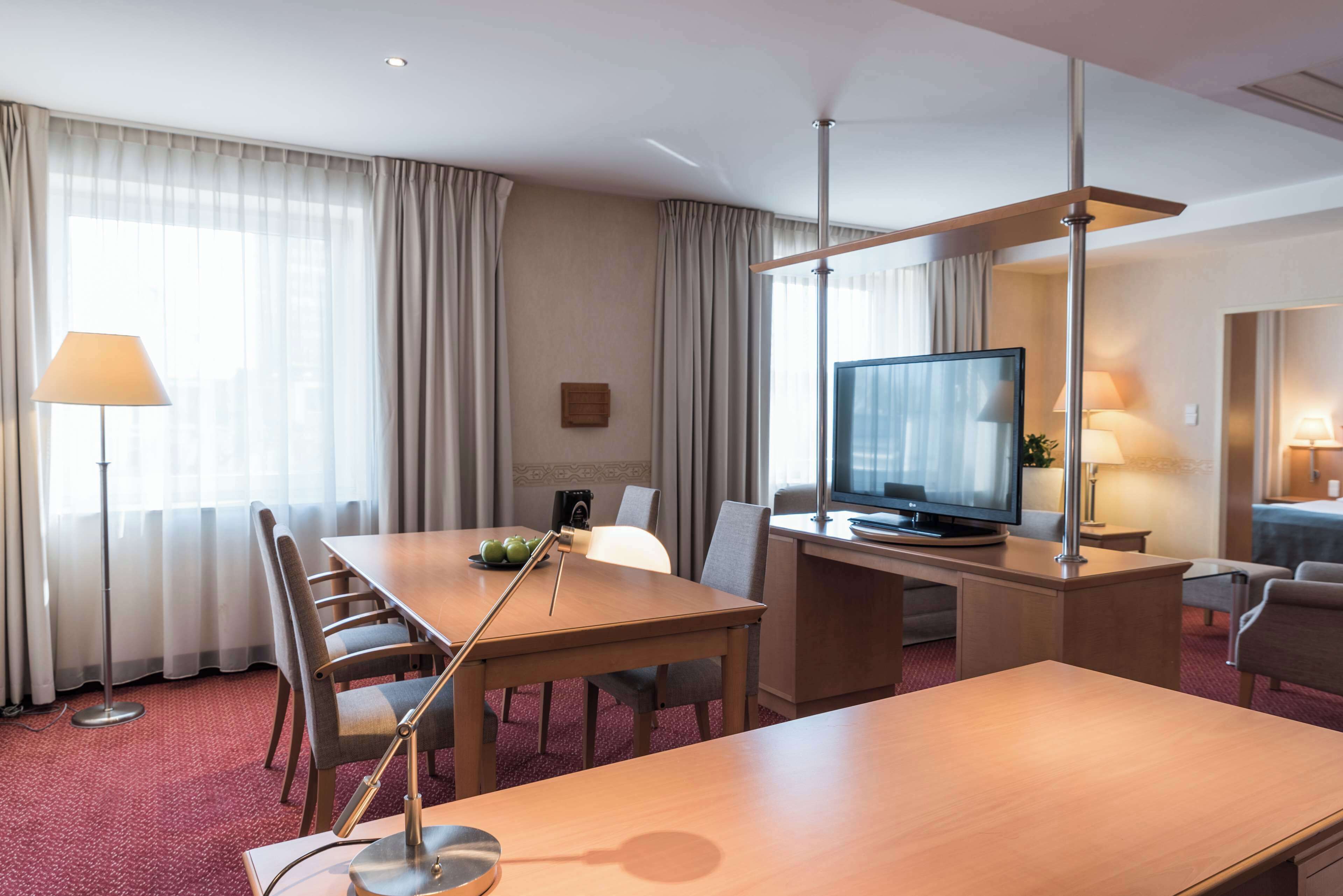 A hotel room with a dining table, chairs, a television, a desk, and a lamp.