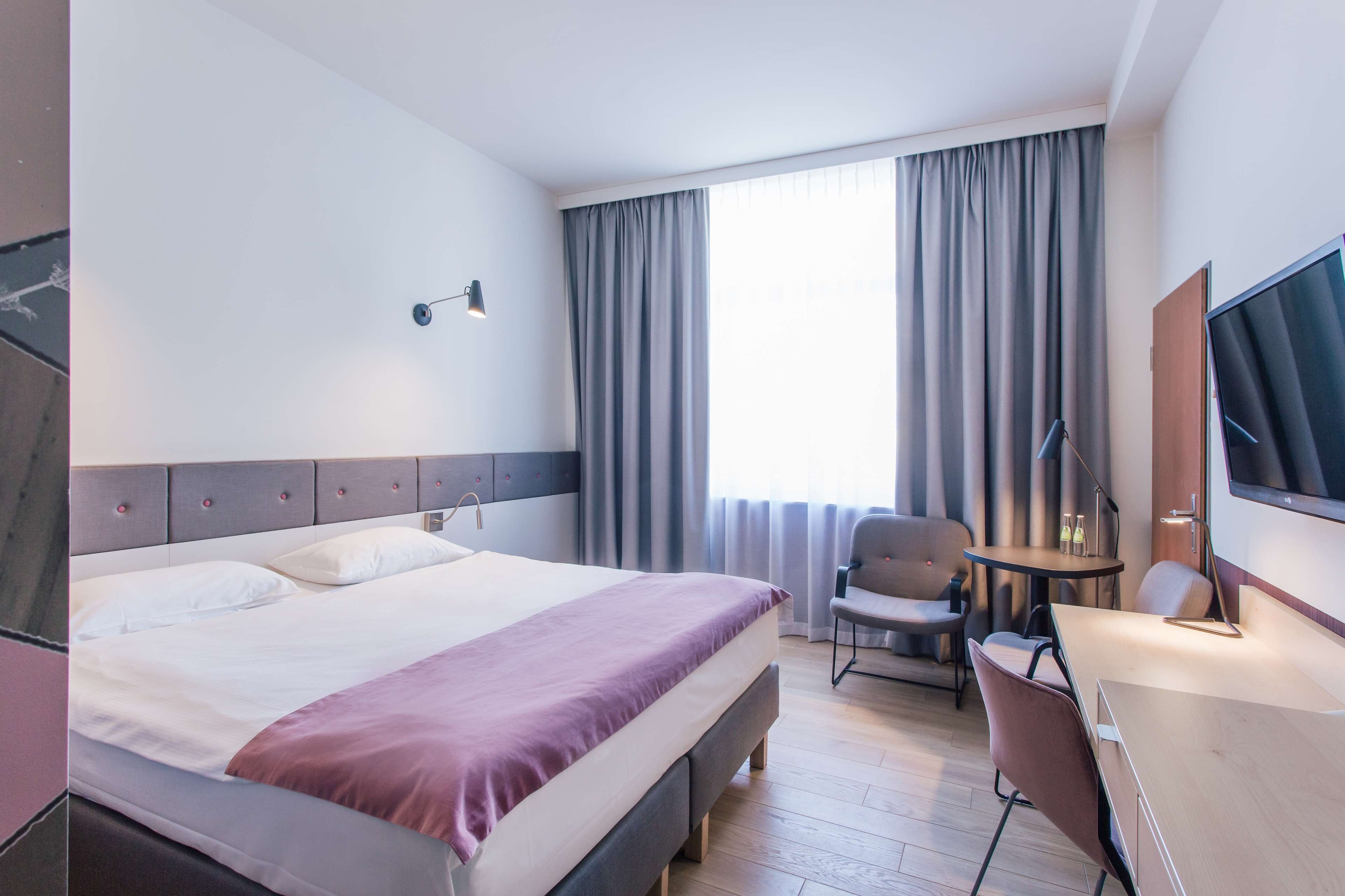 Une chambre d’hôtel avec un grand lit, un bureau, une chaise, une télévision et une fenêtre avec des rideaux.
