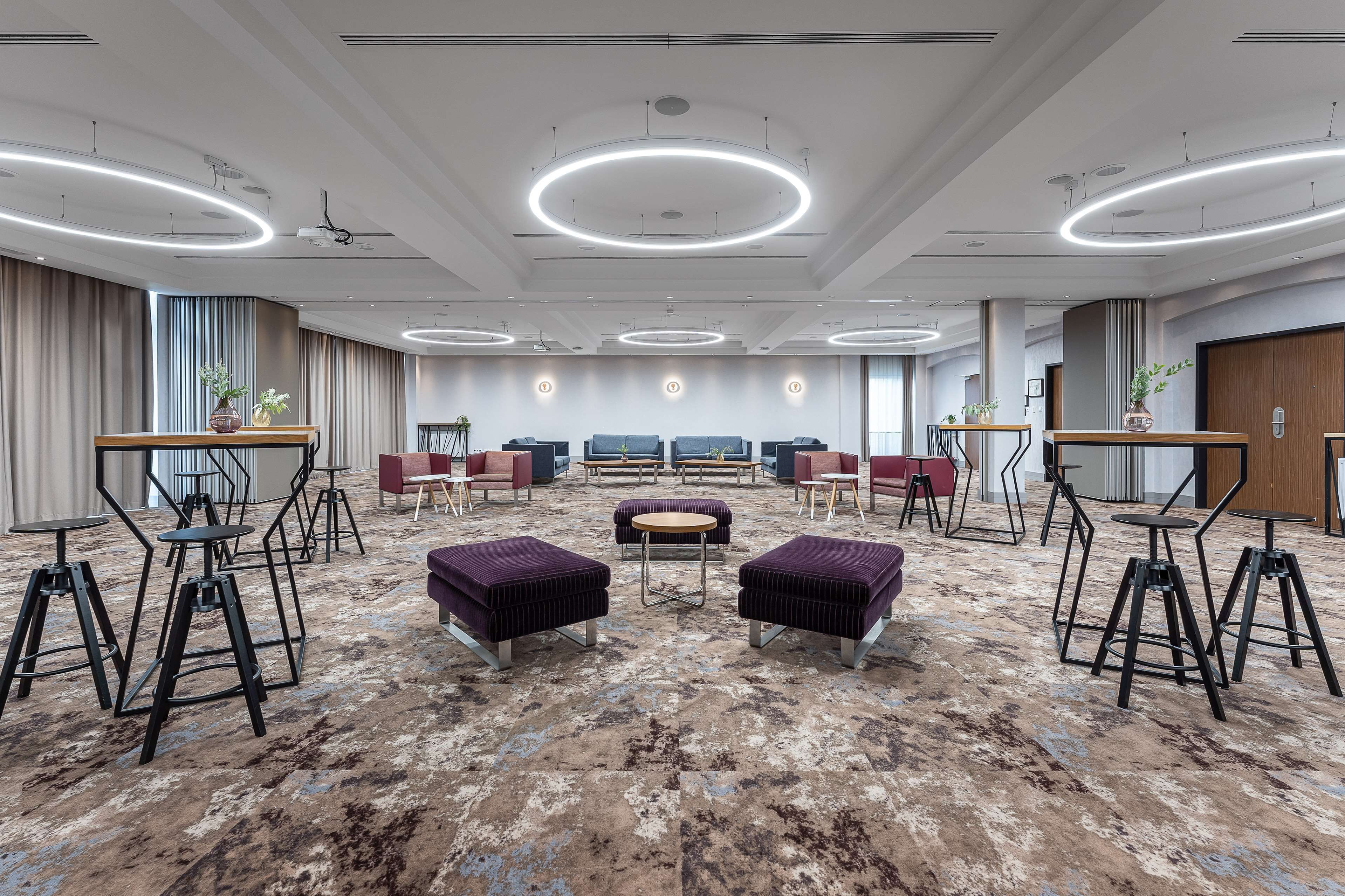 Un hall d’hôtel spacieux au design moderne, comprenant un espace salon central avec des tables et des chaises rondes, une moquette à motifs et un plafond doté de luminaires circulaires.