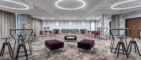 Um amplo hall de hotel com design moderno, destacando uma área de estar central com mesas e cadeiras redondas, um carpete estampado e um teto com luminárias circulares.