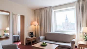 A hotel room with a sofa, a small table with apples, a lamp, and a view of a cityscape through a large window.