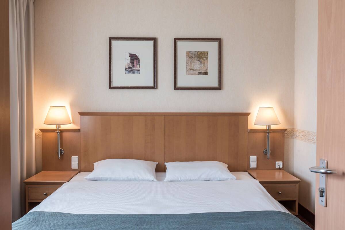 A hotel room with a wooden headboard, two bedside lamps, a bed with white linens, and two framed pictures on the wall.