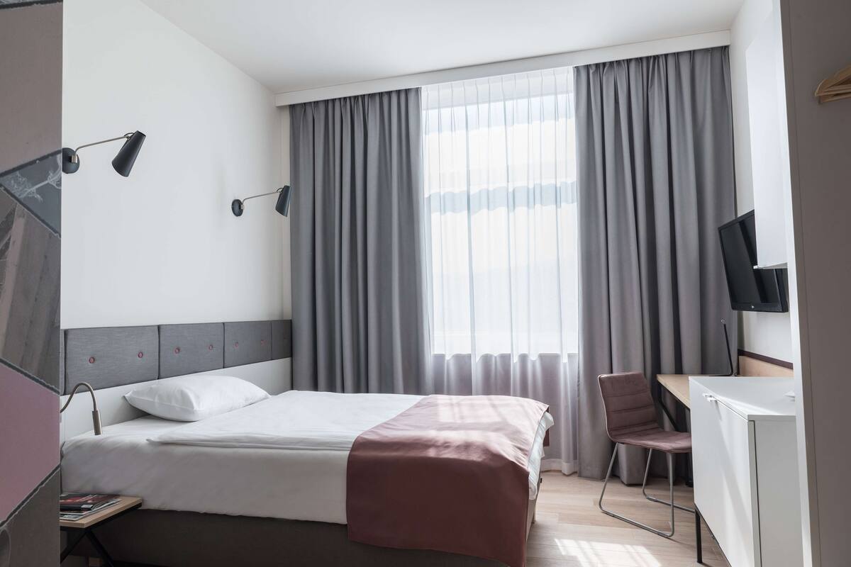 A hotel room with a bed, a desk with a chair, a TV, and a window with curtains.