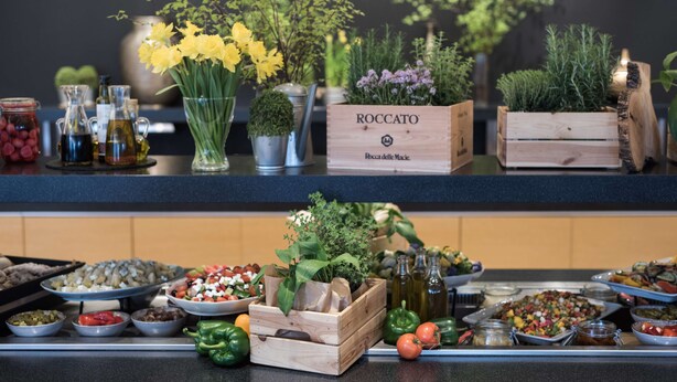 Una mesa de buffet con diferentes alimentos, una caja de madera con el rótulo 'ROCCATO' y plantas decorativas.