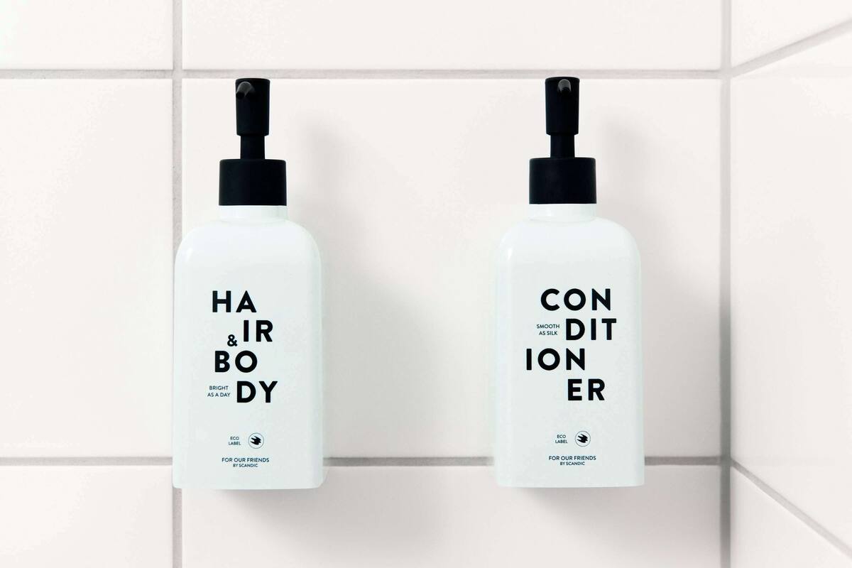 Two white bottles with black pumps, labeled "HAIR & BODY" and "CONDITIONER," hanging on a white tiled wall.