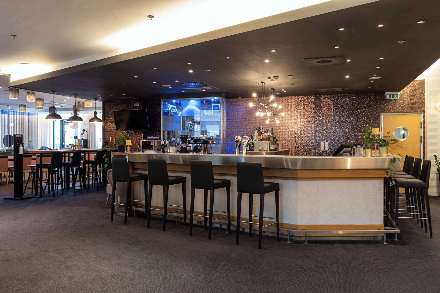 A modern bar area with a bar counter, multiple stools, a television screen, and a decorative wall with a mosaic pattern.
