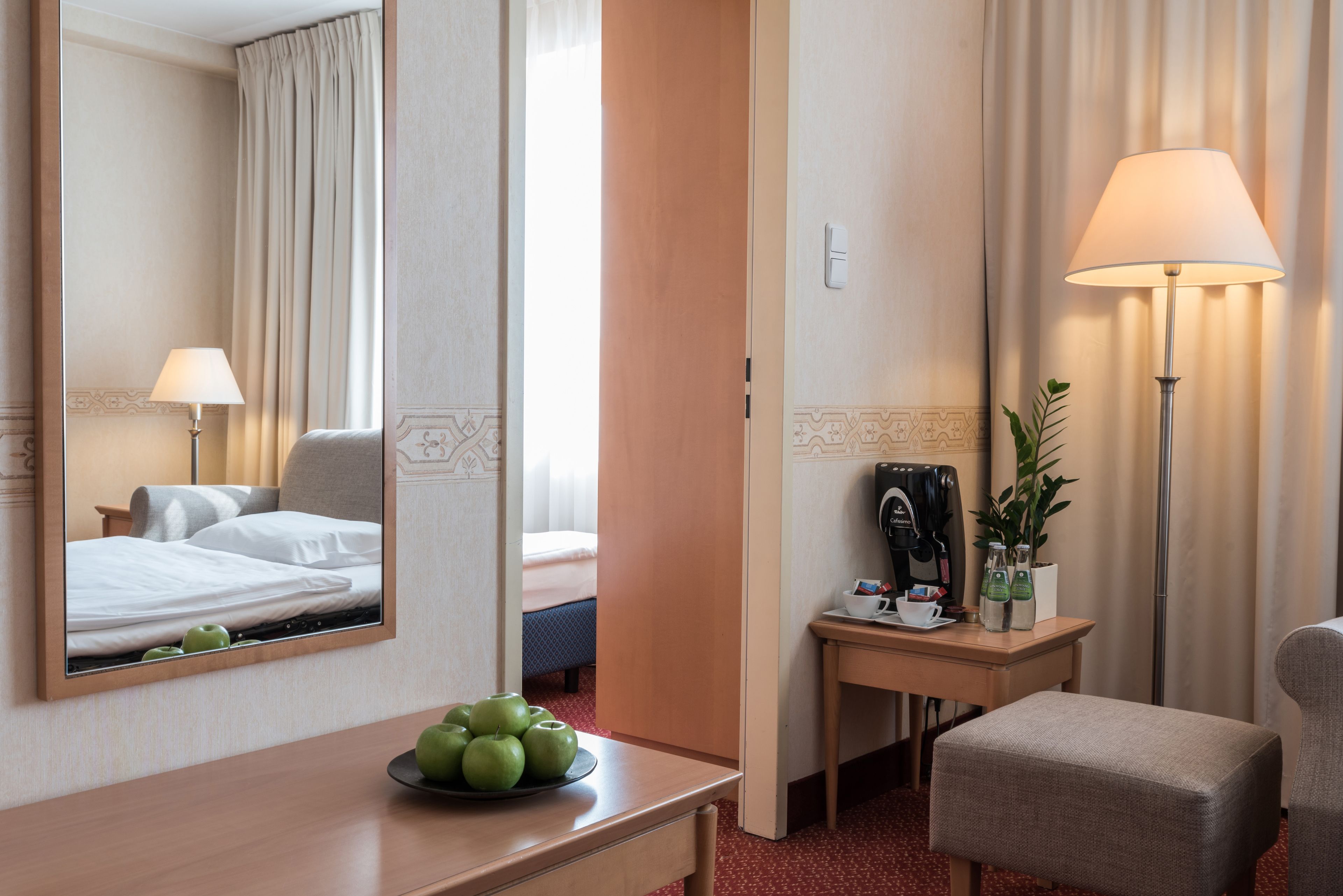 A hotel room with a bed, a desk with a lamp, a chair, a small table with apples, and a mirror reflecting the room.