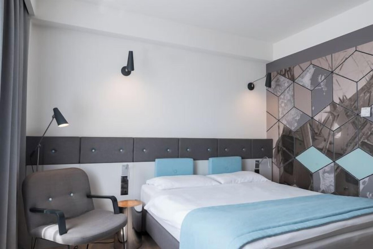 A hotel room with a bed, a chair, a desk, and a wall with a geometric pattern.