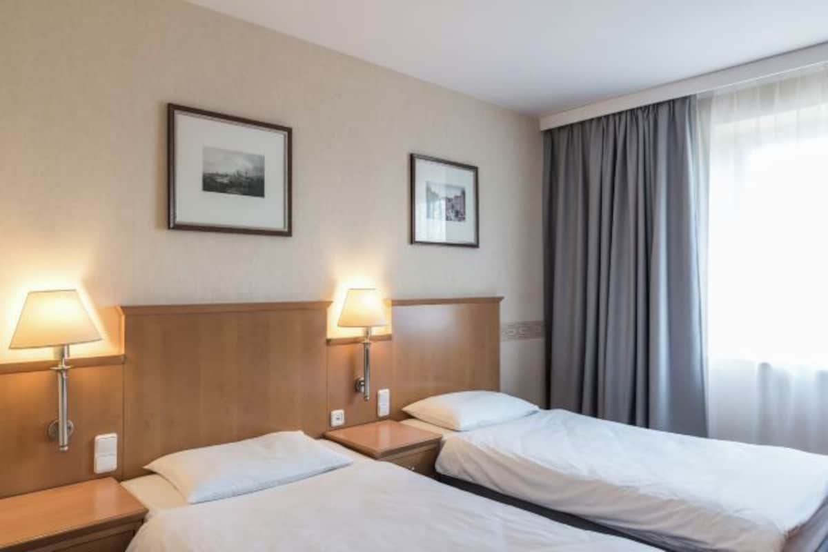 Habitación de hotel con dos camas, cada una con mesita de noche y lámpara, cuadros en la pared y una ventana con cortinas.