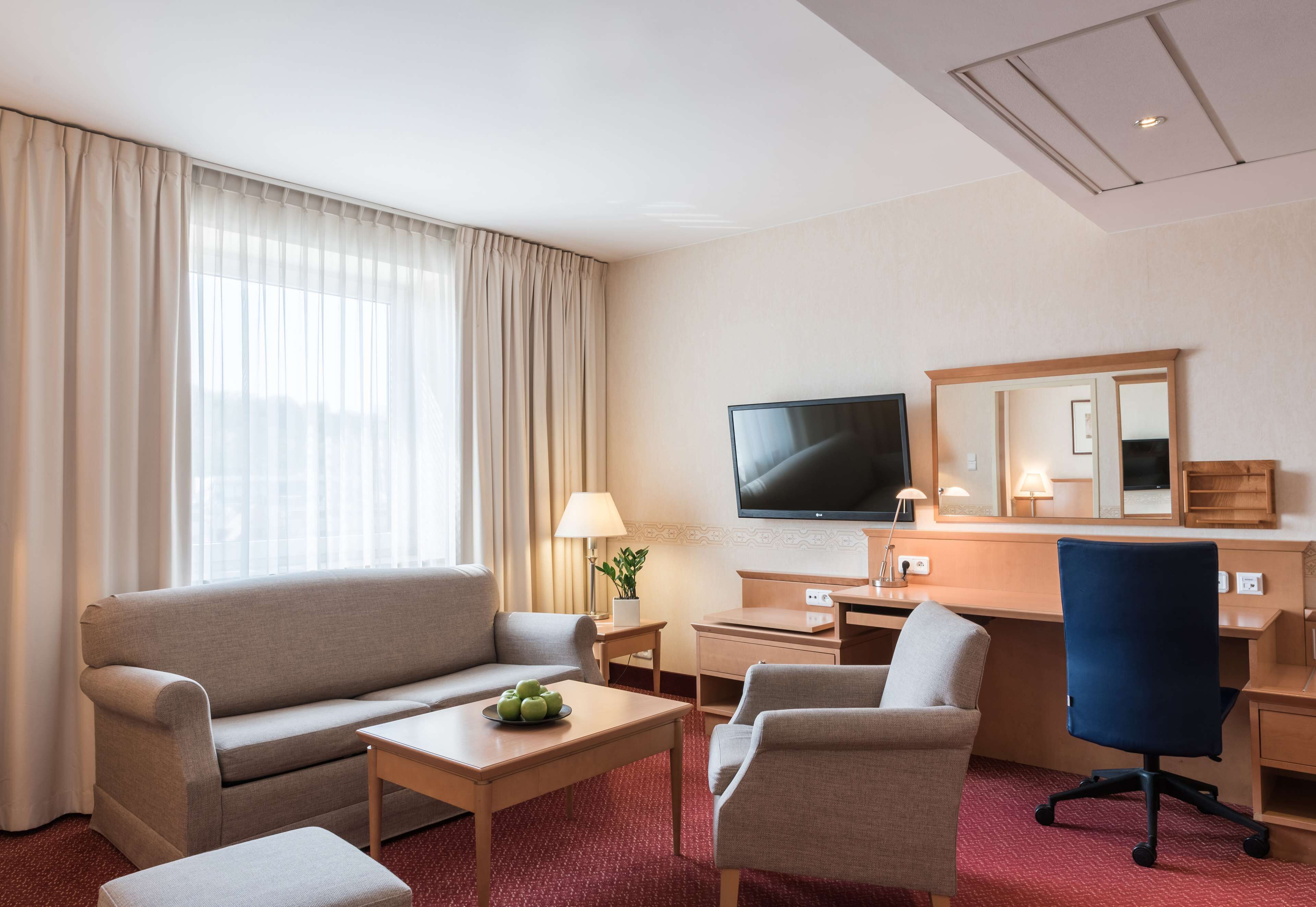 A hotel room with a sofa, a desk, a chair, a television, and a large window with curtains.