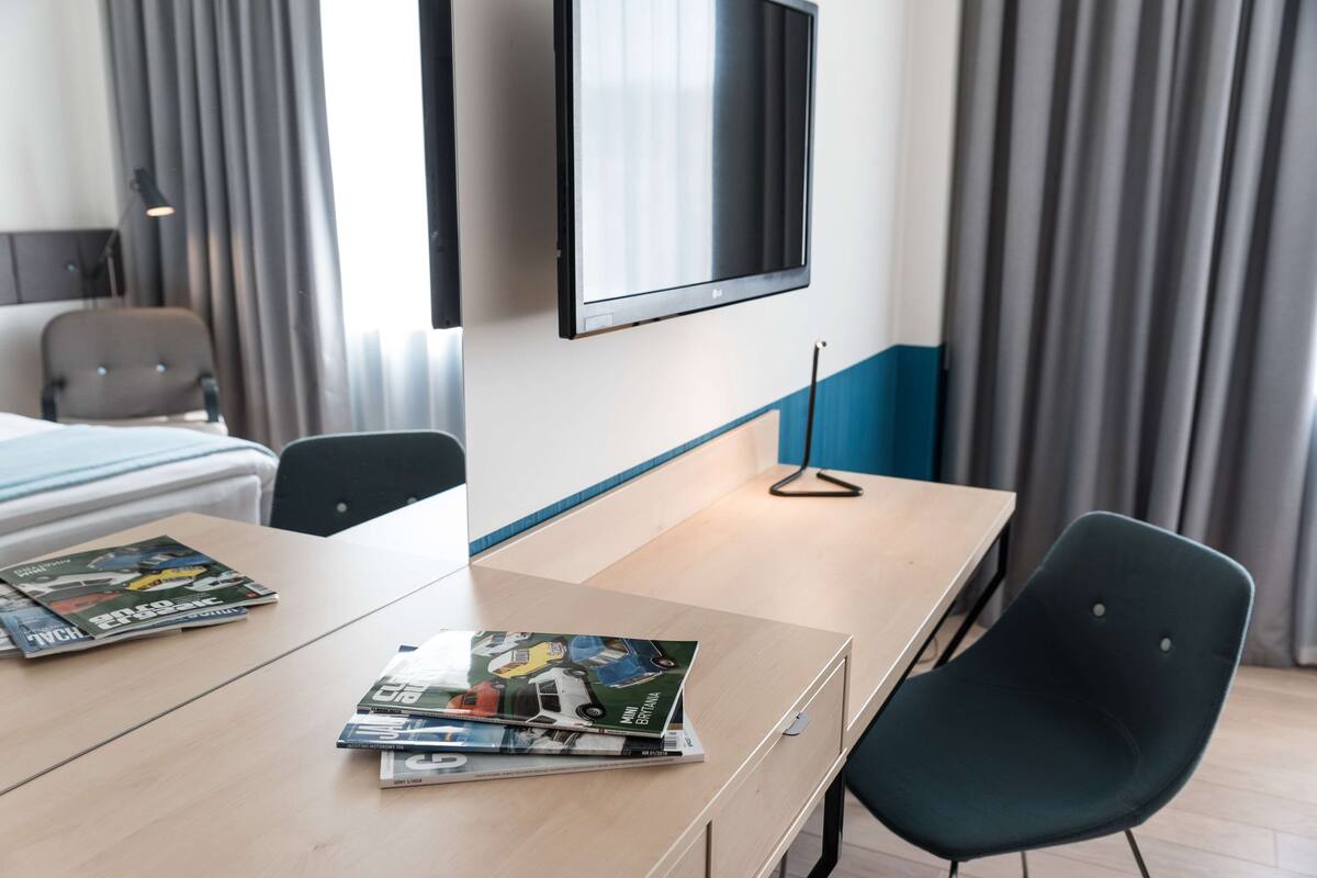 A hotel room with a bed, a TV mounted on the wall, a wooden desk with magazines, and a chair.