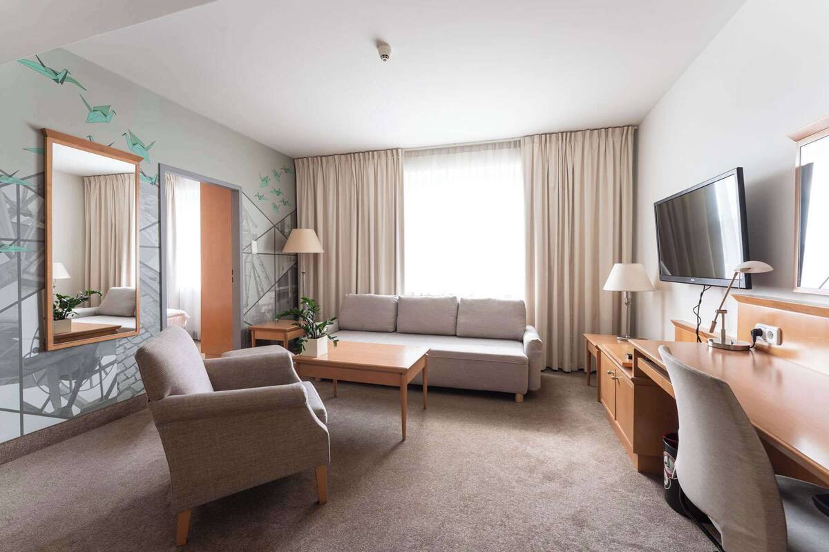 A hotel room with a sofa, armchair, desk, TV, and a large window with curtains.