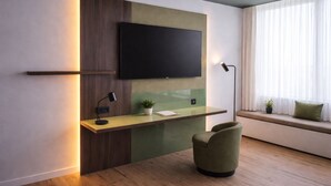 A modern living room with a flat-screen TV mounted on a wall, a wooden desk with a lamp, a green chair, and a window with curtains.