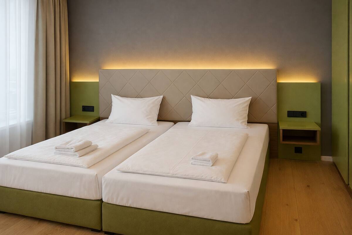 Cama doble con dos almohadas y toallas dobladas, mesitas de noche verdes y cabecero de color claro.