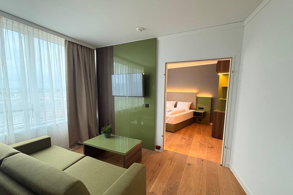 Una habitación de hotel moderna con una pared verde de acento, un televisor de pantalla plana sobre la cama, una mesita de noche pequeña y vistas de la ciudad a través de la ventana.