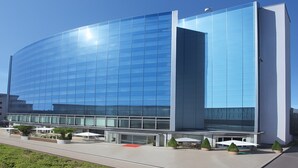 A modern glass building with a curved facade, outdoor seating with umbrellas, and a clear sky.
