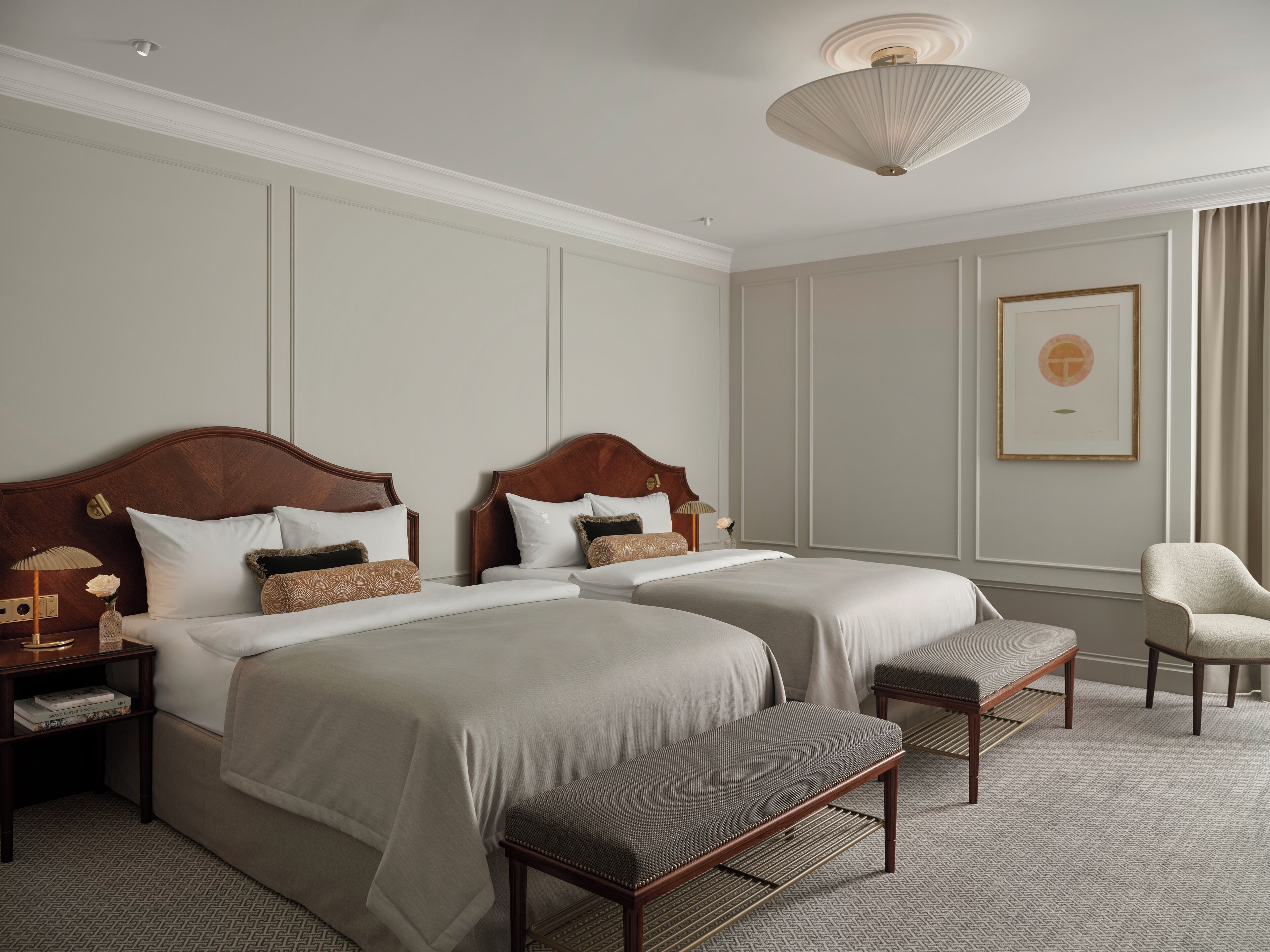 Habitación de hotel con dos camas, un banco, una silla, una lámpara y un cuadro en la pared.