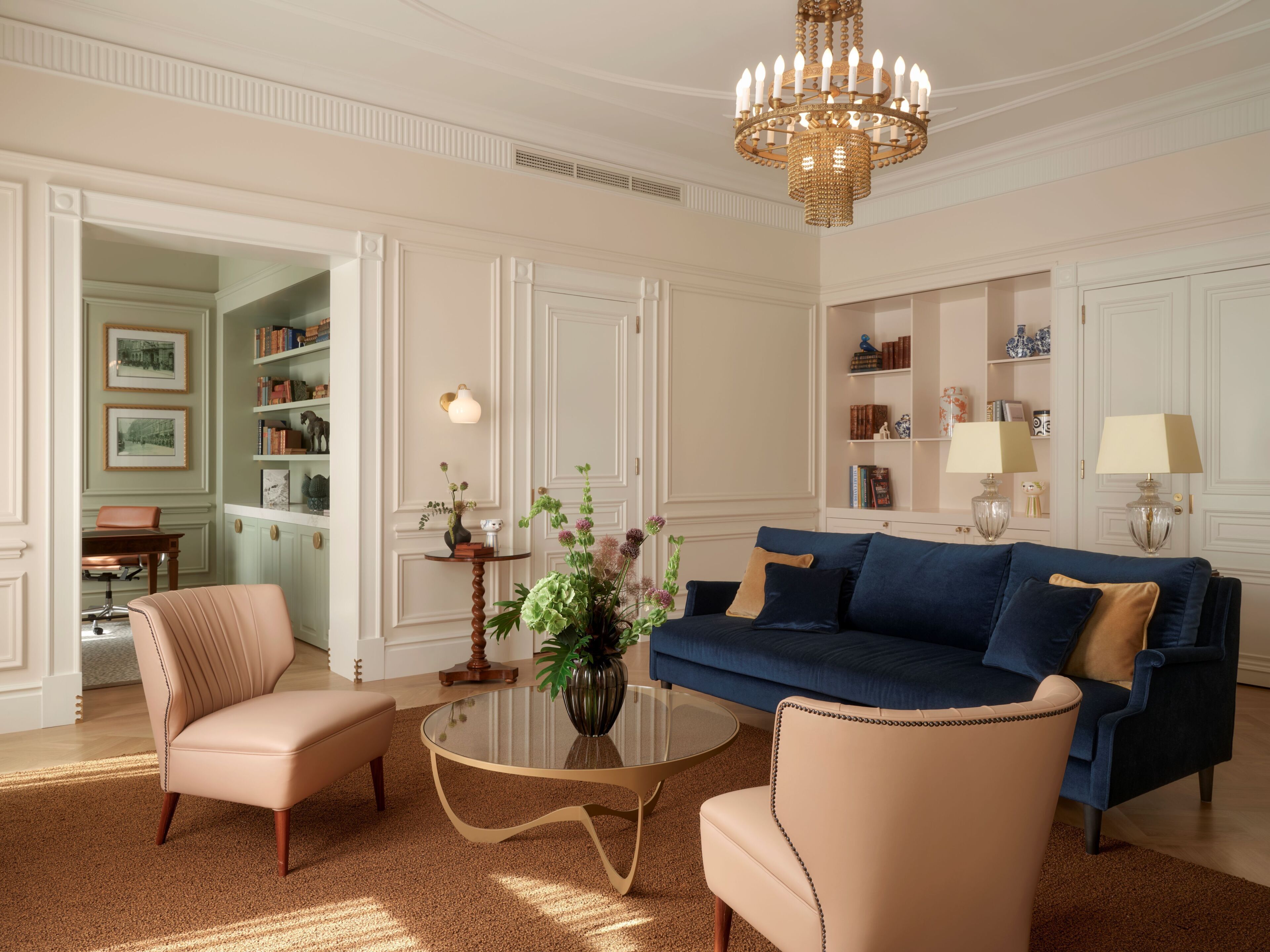 A well-lit living room with a blue sofa, beige armchairs, a glass coffee table, and a decorative chandelier.