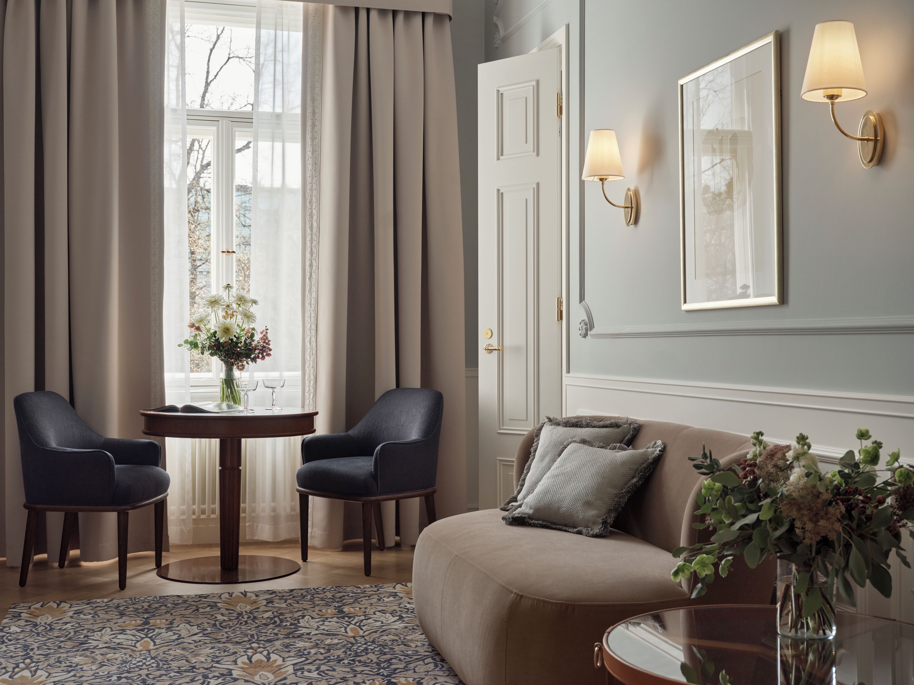 A room with a beige sofa, two armchairs, a round table with a vase of flowers, and a large window with curtains.