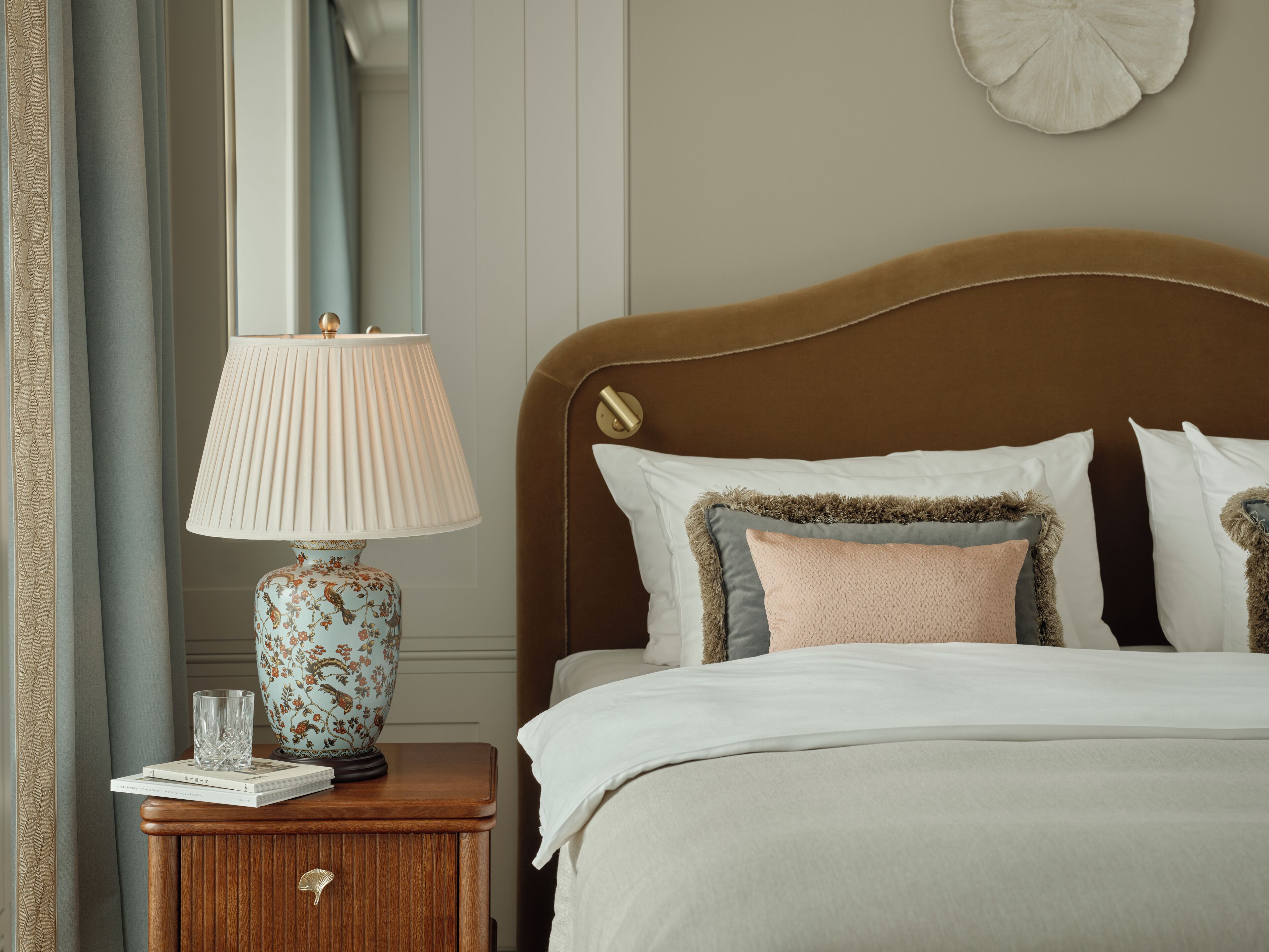 A neatly made bed with white linens, a wooden nightstand with a floral lamp, and a wall-mounted decorative piece.