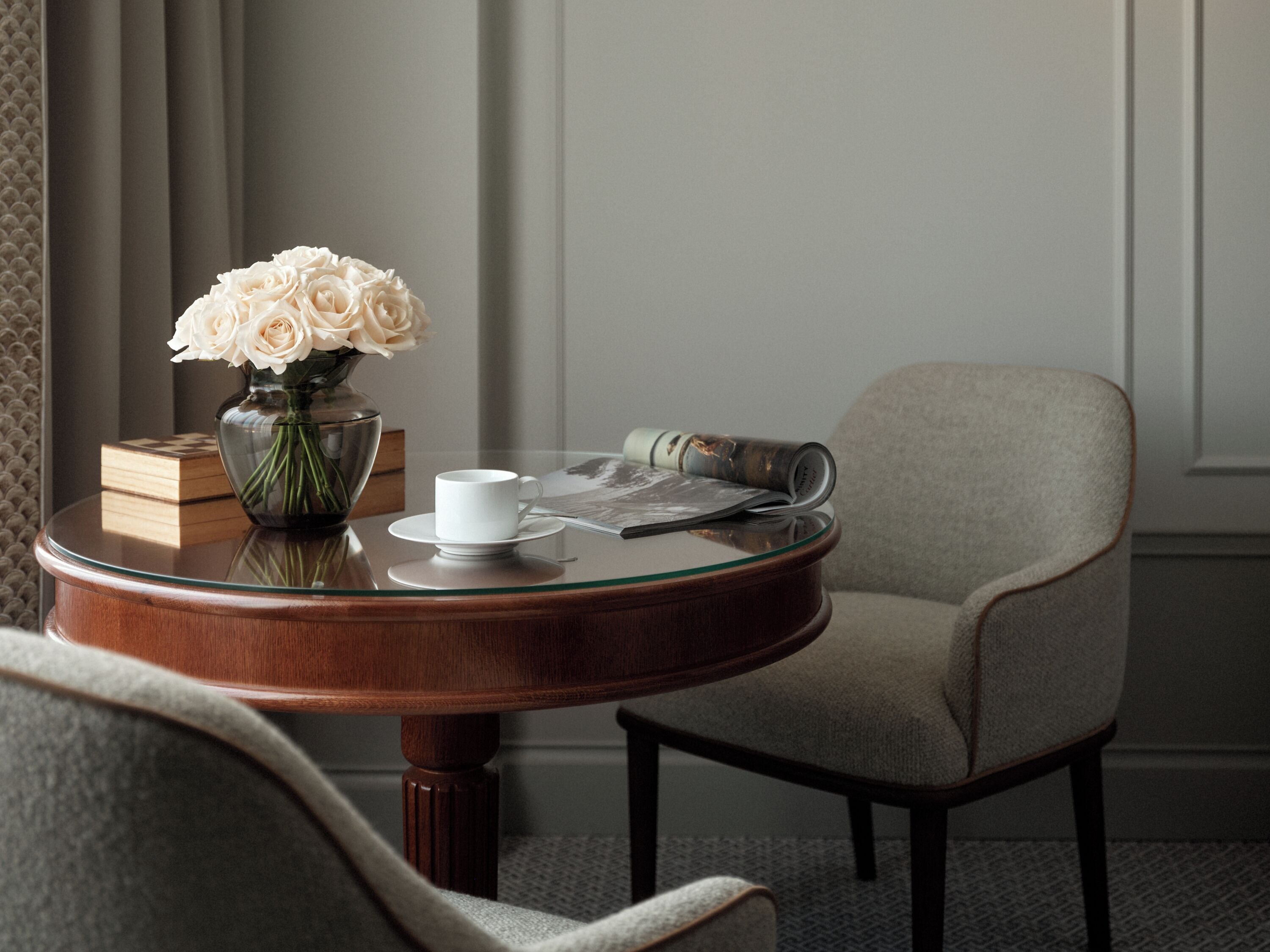 A round wooden table with a glass top, a white cup, a vase with white flowers, and a rolled magazine.
