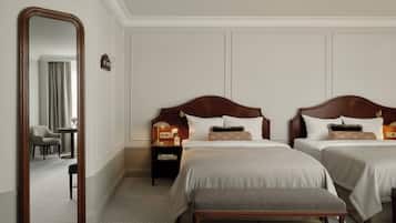 A hotel room with two beds, a large mirror, a bench, a nightstand with a lamp, and a view of a hallway with a chair and curtains.