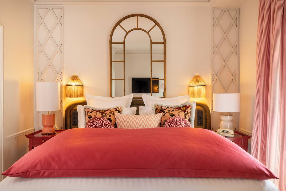 A neatly made bed with a red bedspread, flanked by two bedside tables with lamps, and a large mirror above the bed.