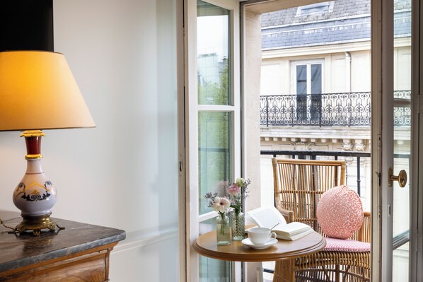 A room with a table, a lamp, a chair, a vase with flowers, and a view of a balcony with a railing. - Hôtel Le Lavoisier & Spa (Paris)