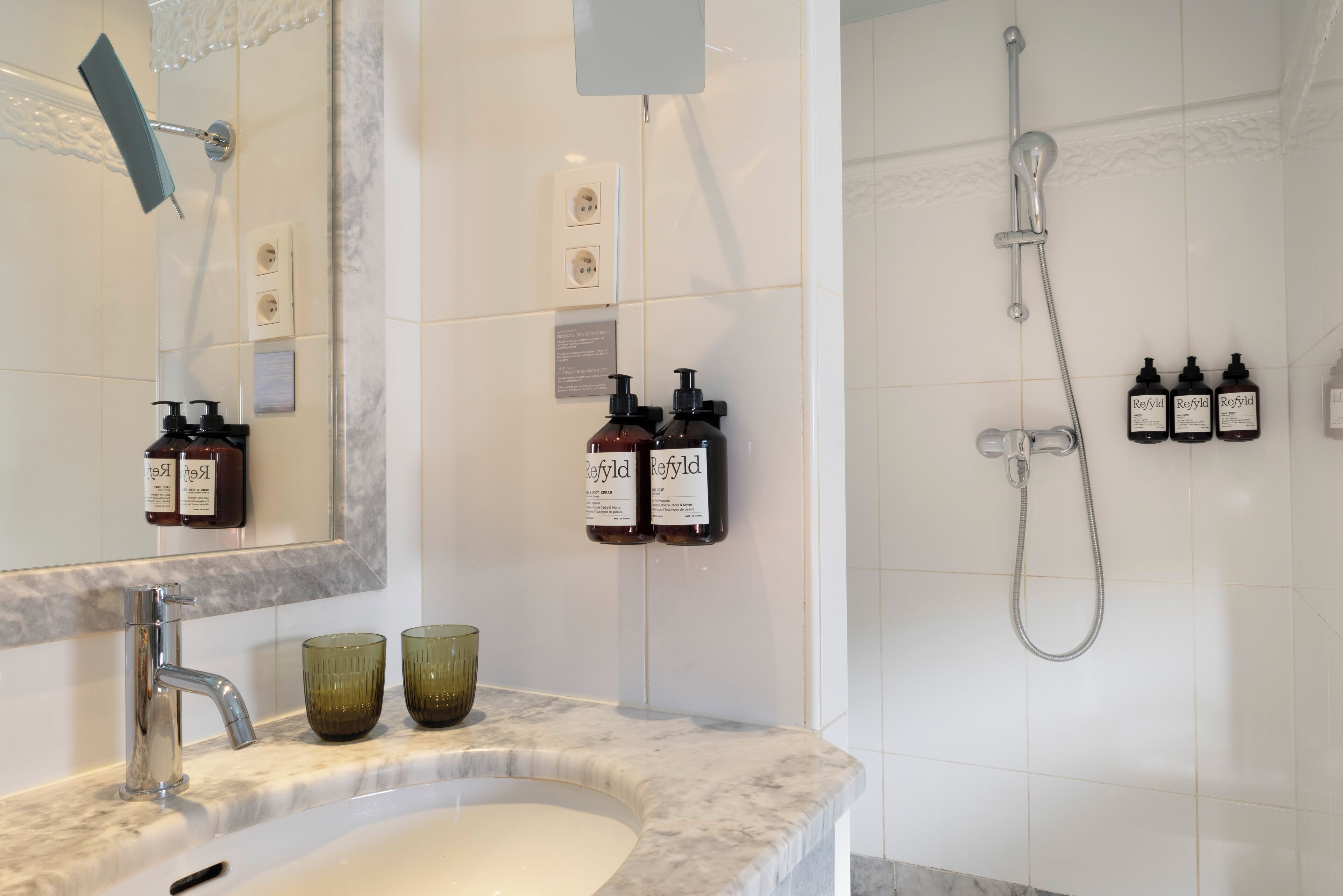 A bathroom with a sink, mirror, and shower. There are bottles of Refyld body cream and soap on the wall.