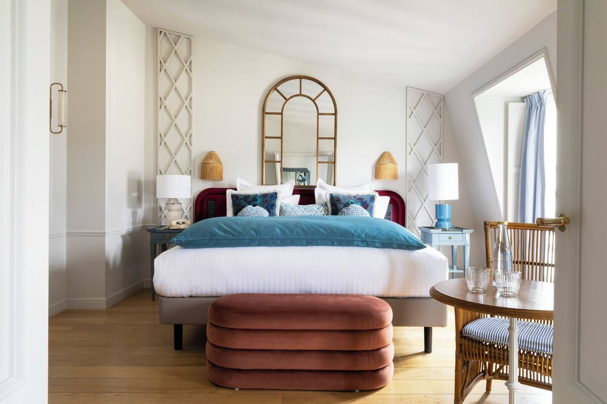 A bedroom with a large bed, a round table with chairs, a mirror, and a window with curtains.