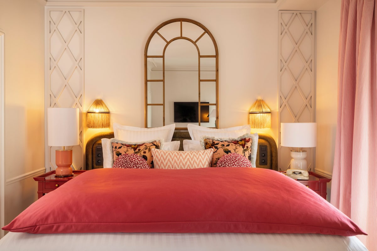 A neatly arranged bed with a red bedspread, flanked by two bedside tables with lamps, a large mirror above the bed, and a decorative wall panel.