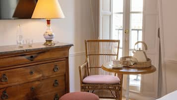 A room with a wooden dresser, a wicker chair, a small round table with a lamp, and a window with curtains.