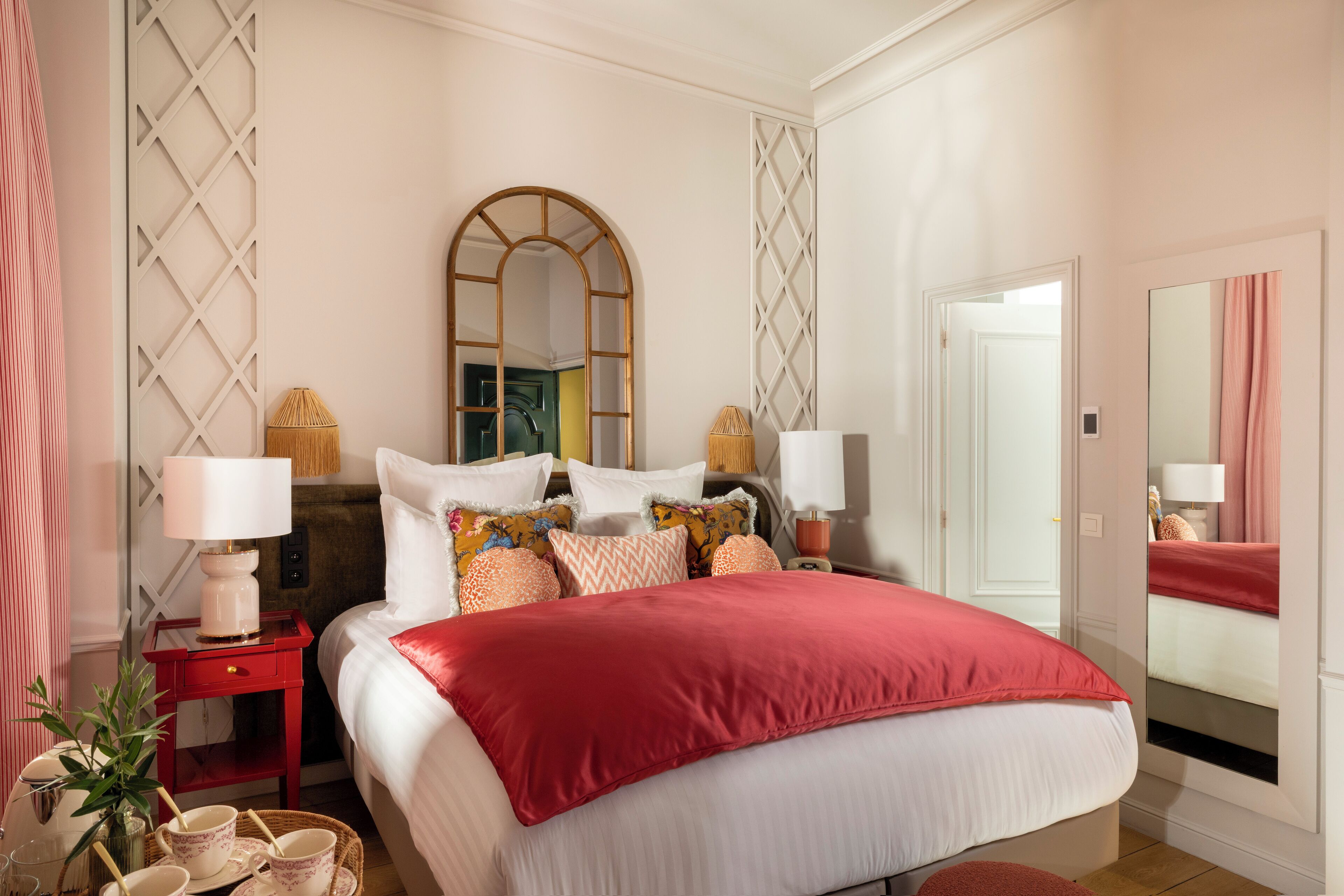 A neatly arranged bedroom with a large bed, a bedside table with a lamp, a mirror, and a small side table with a basket.