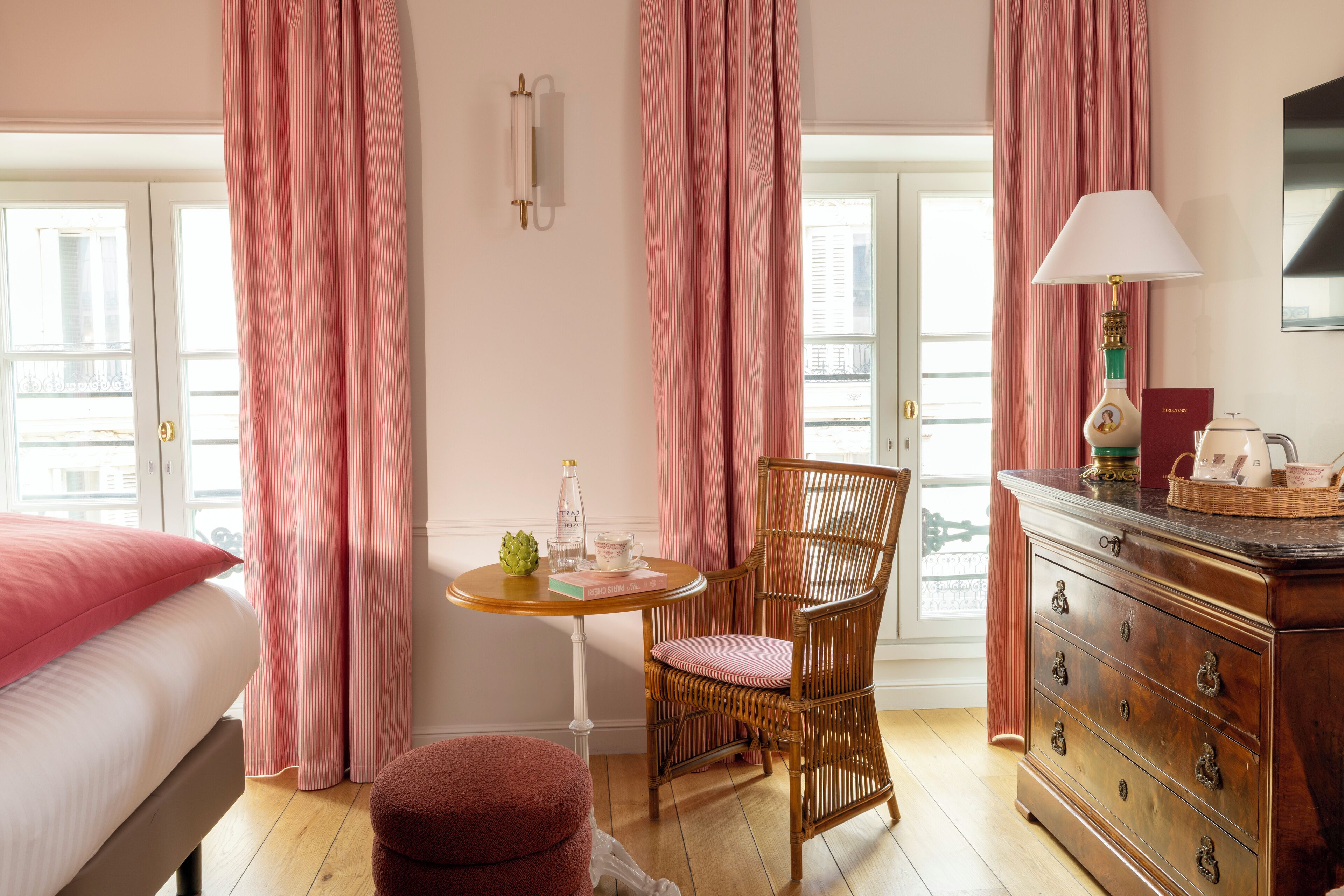 A hotel room with a bed, a wooden dresser, a wicker chair, a small round table with a bottle and books, and pink curtains.