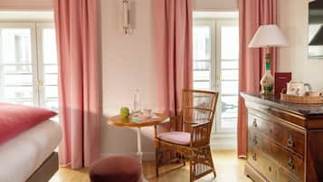A hotel room with a bed, a wooden dresser, a wicker chair, a small round table with a bottle and books, and pink curtains.