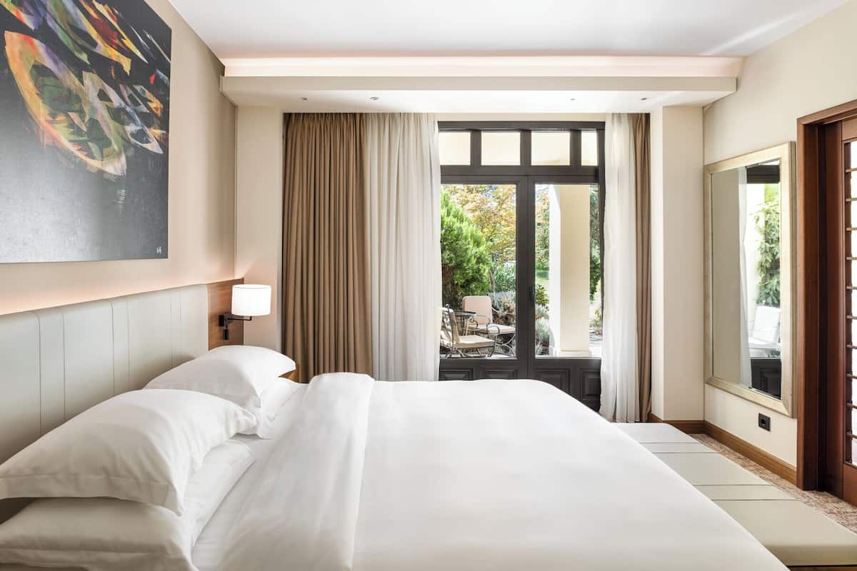 A hotel room with a large bed, a nightstand with a lamp, a view of the garden through a window, and a painting on the wall.