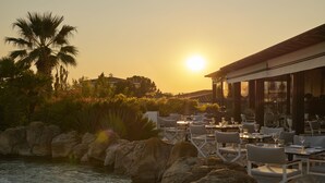 A restaurant with outdoor seating overlooking a body of water, palm trees, and a sunset. - Hyatt Regency Thessaloniki (Thermi)
