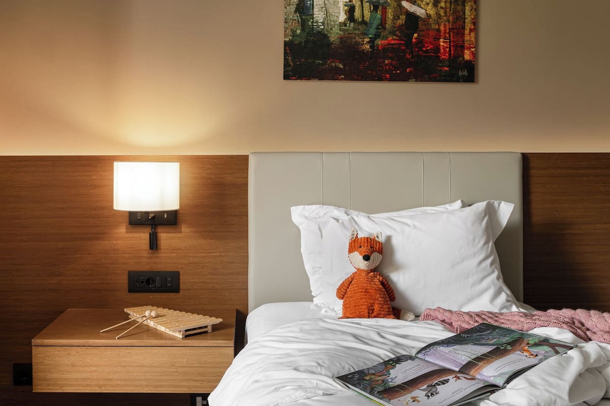 A hotel room with a neatly made bed, a nightstand with a lamp, a painting on the wall, and a plush toy on the bed.