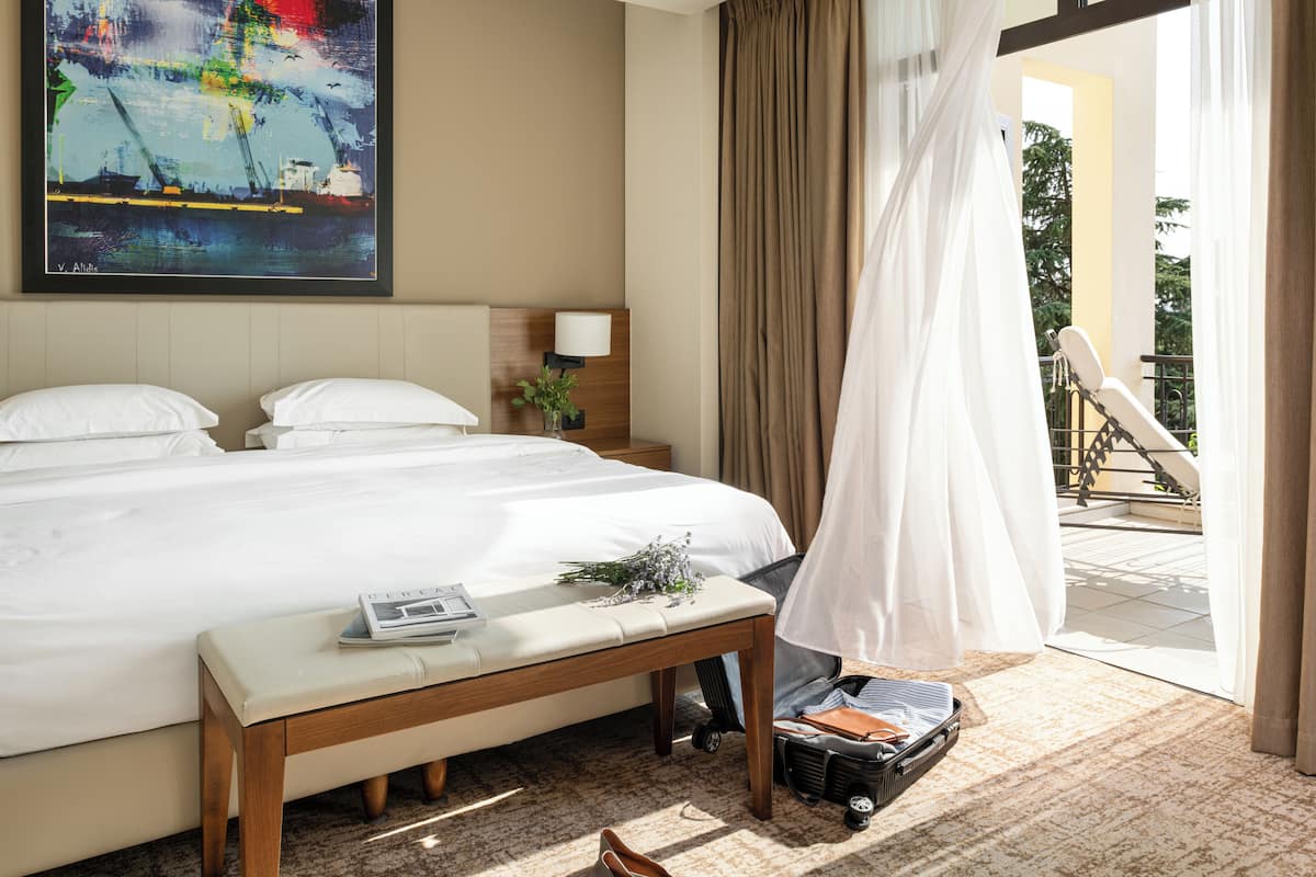 A hotel room with a large bed, a bedside table, a suitcase, and a painting on the wall.