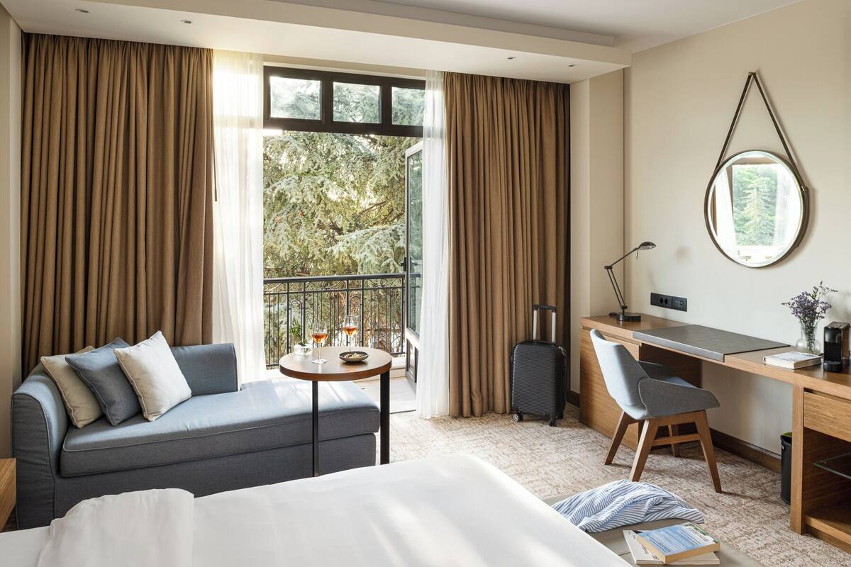 A hotel room with a bed, a desk, a chair, a small table with glasses of wine, and a view of trees through large windows.
