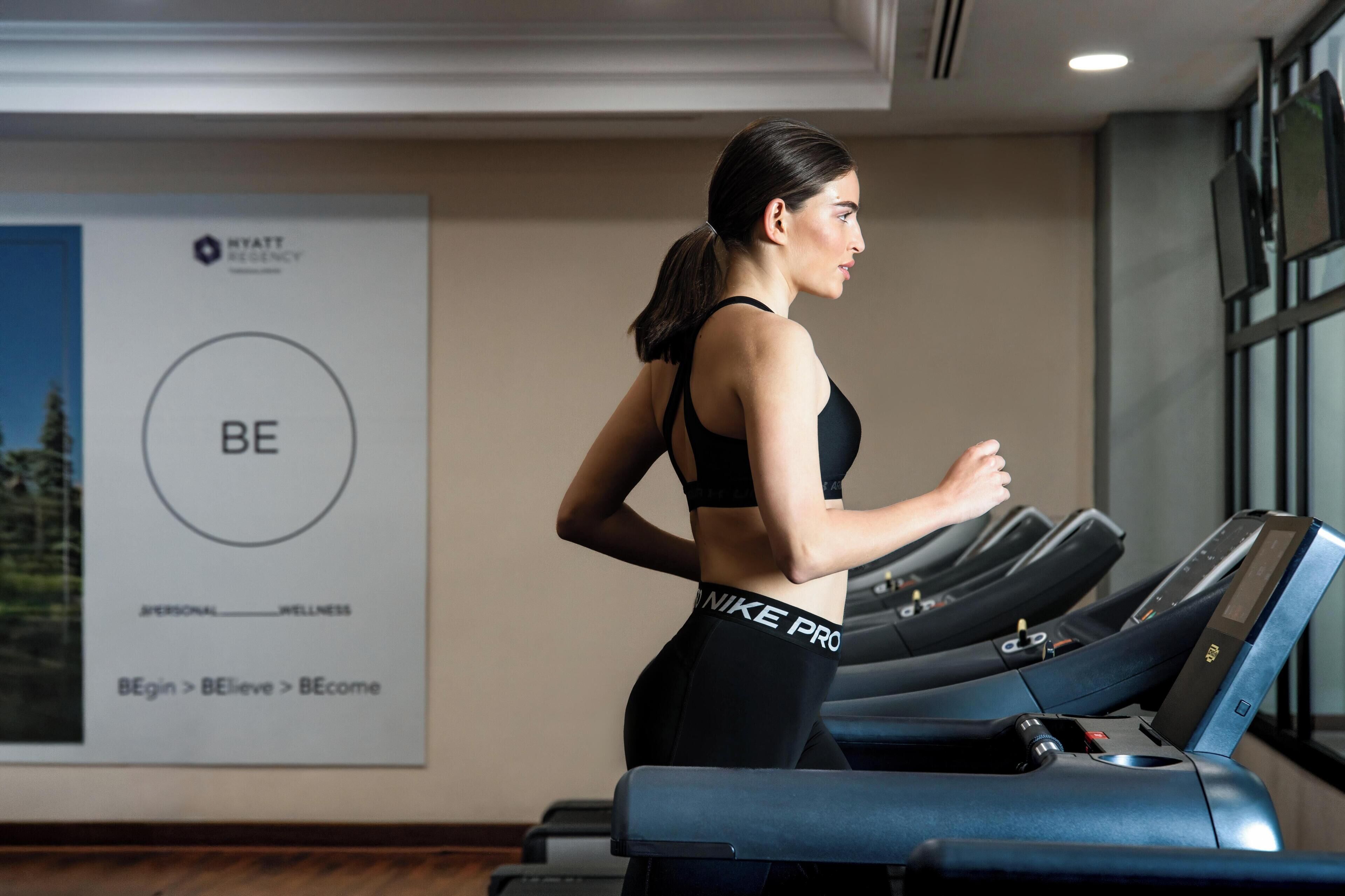 A woman running on a treadmill in a gym with a poster on the wall that reads "BE" and "NIKE PRO".