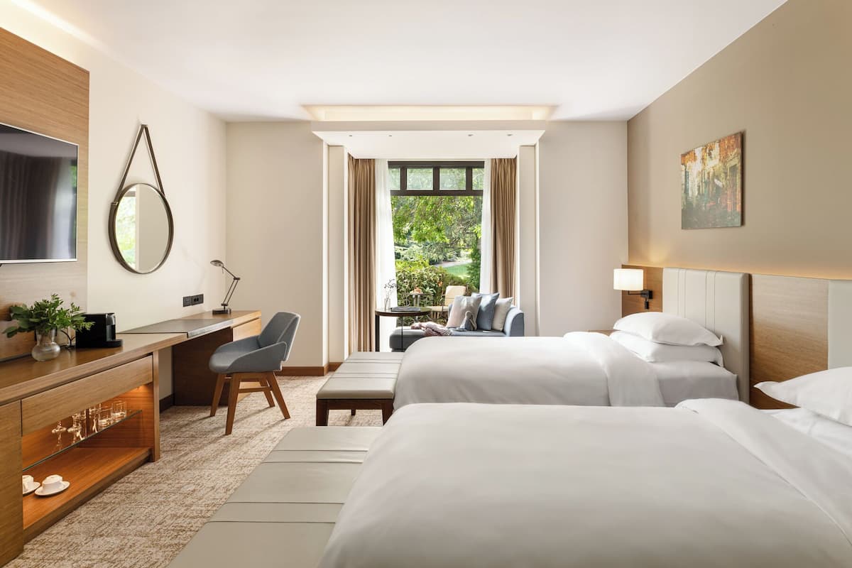 A hotel room with a desk, two beds, a TV, and a view of greenery.
