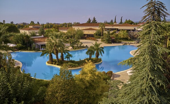 Una amplia piscina curvada rodeada de palmeras y follaje, con edificios al fondo.
