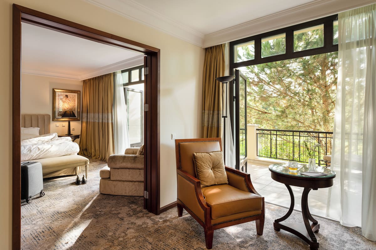 A hotel room with a large window, a leather armchair, a bed, a small table, and a luggage trolley.