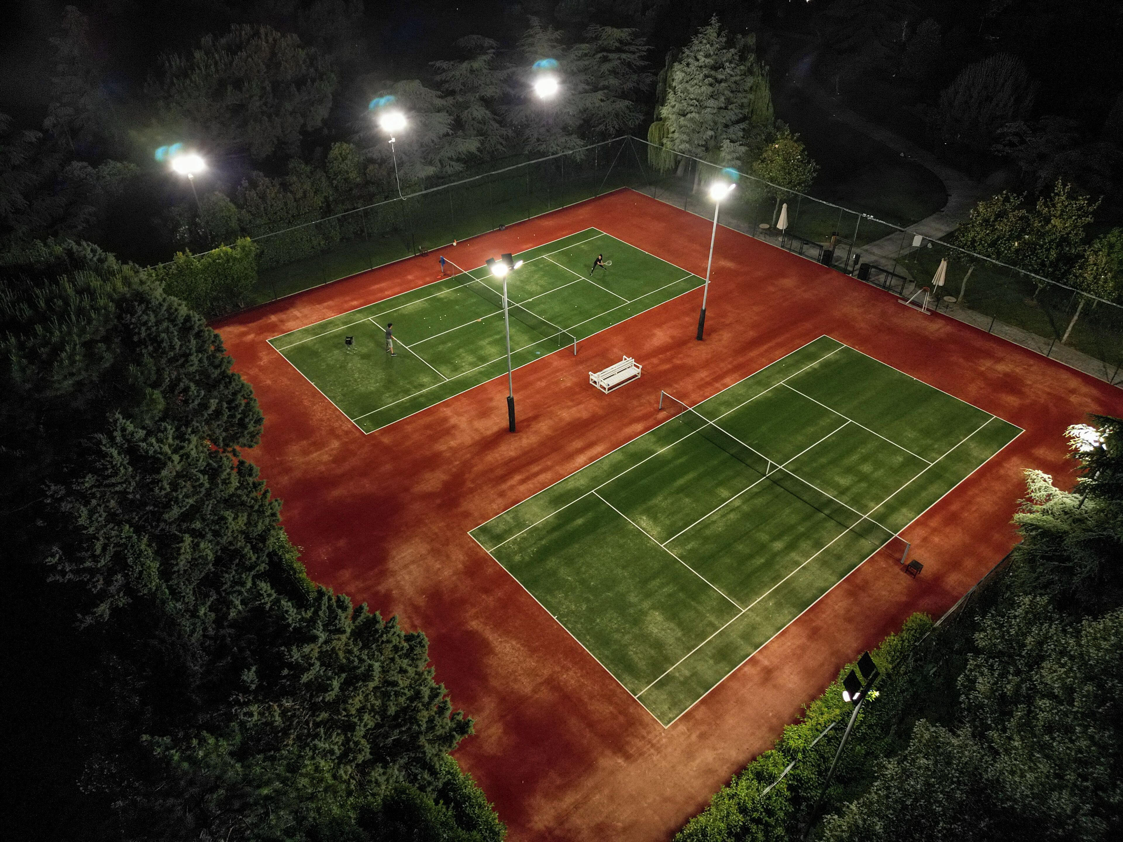 En udendørs tennisbane omgivet af træer, med flere lygtepæle der giver lys.